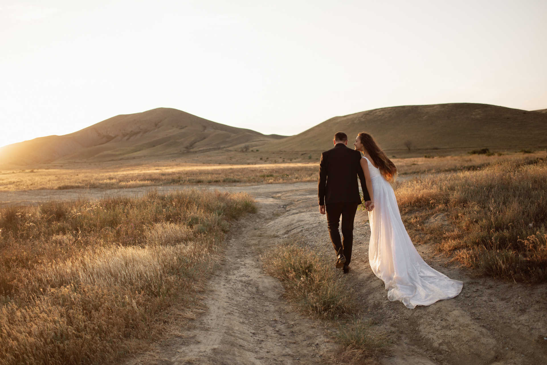 Wedding couple holding hands walks along a path in the field under the sunset mountains in a golden light. Bride with luxury flowing hem walks in safari holding hand of her lover and smiling.